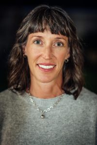 Headshot of Dr. Jennifer Watt wearing light gray sweater with silver necklace. Dr. Watt has bangs and shoulder-length wavy brown hair.
